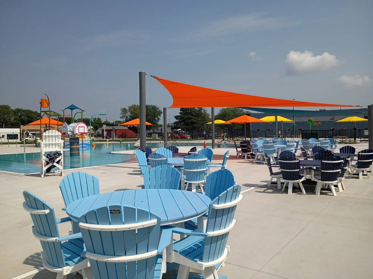 Outdoor poolside patio area with light blue Adirondack-style tables and chairs under large orange shade sails, with a swimming pool, lifeguard chair, and playground visible in the background.